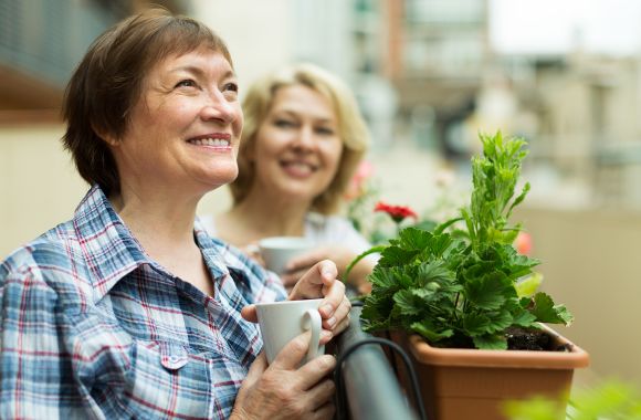 Zwei Frauen stehen lächelnd auf dem Balkon, während sie Tassen halten.