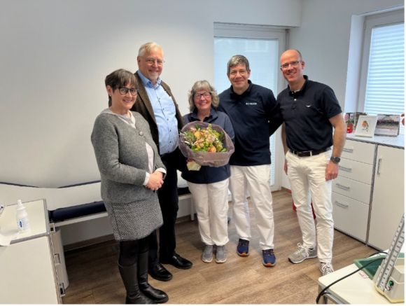 Fünf Personen in einem Behandlungsraum posieren lächelnd mit einem Blumenstrauß. Auf dem Bild zu sehen: Anja Herder-Peyrounette, Dr. Harald Menker, Dorothee Massing,  Dr. Stergios Tsolakidis, Dr. Wilhelm Litz