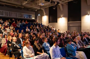 Ein Publikum sitzt aufmerksam in einem Veranstaltungsraum und verfolgt eine Präsentation auf der Bühne.