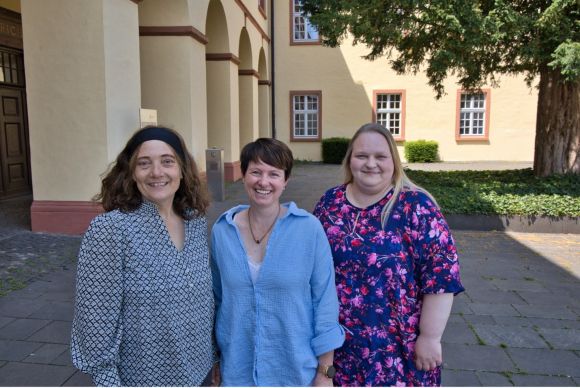 Drei Frauen, die Demenzlotsinnen, lächeln fröhlich vor einer Gebäude-Fassade im Freien.
