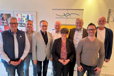 Eine Gruppe von acht Personen steht lächelnd vor einem Banner der Gesundheitsregion Siegerland.