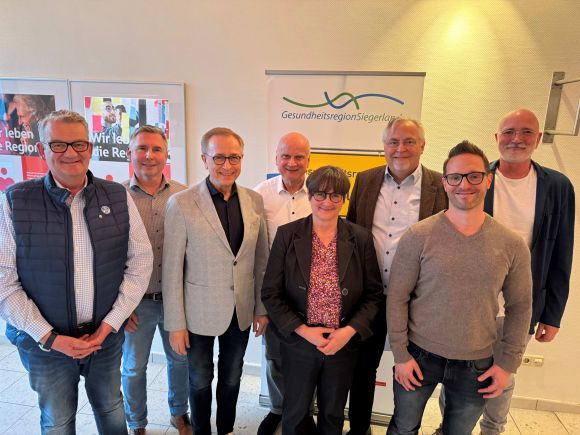 Eine Gruppe von acht Personen steht lächelnd vor einem Banner der Gesundheitsregion Siegerland.