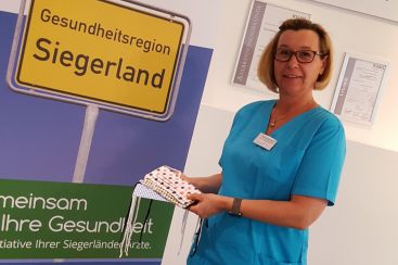 Eine Frau in blauem Kasak hält mehrere Mund-Nasen-Schutzmasken vor einem Schild der Gesundheitsregion Siegerland.