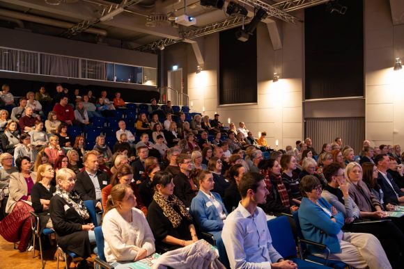 Ein Publikum sitzt aufmerksam in einem Veranstaltungsraum und verfolgt eine Präsentation auf der Bühne.