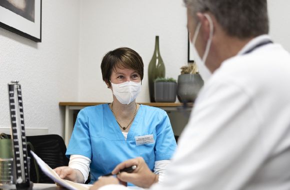 Eine Frau in blauem Kasak lächelt während einer Beratung mit einem Patienten in einem modernen Behandlungsraum.