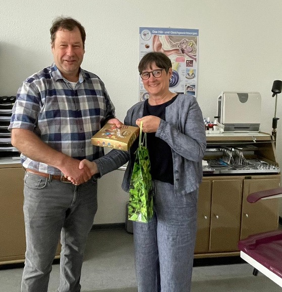 Dr. Alexander Heß und Anja Herder-Peyrounette schütteln sich die Hände, während sie Geschenke in einem Büro überreichen.