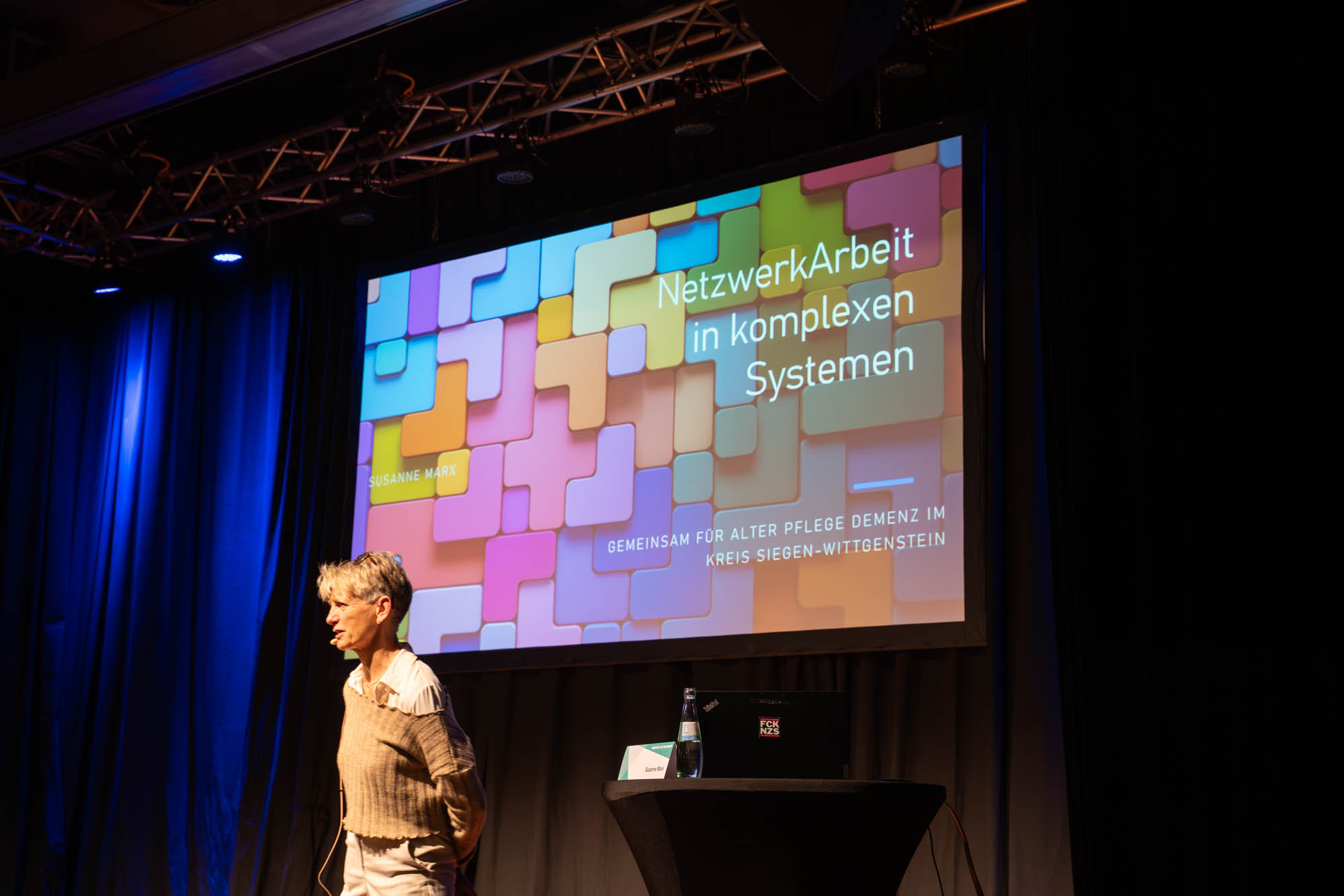 Eine Rednerin präsentiert auf einer Bühne das Thema "NetzwerkArbeit in komplexen Systemen" vor einem Bildschirm.