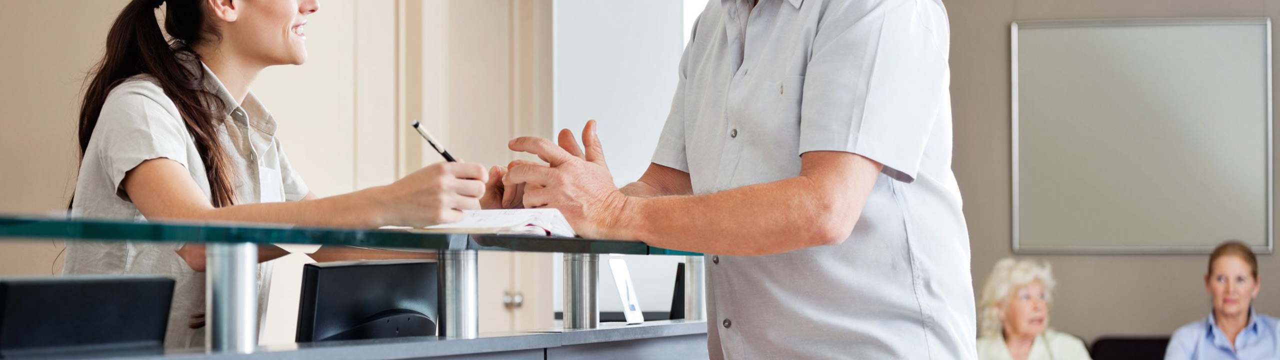 Eine Mitarbeiterin an der Rezeption hilft einem älteren Mann bei einer Anfrage, während andere Patienten warten.