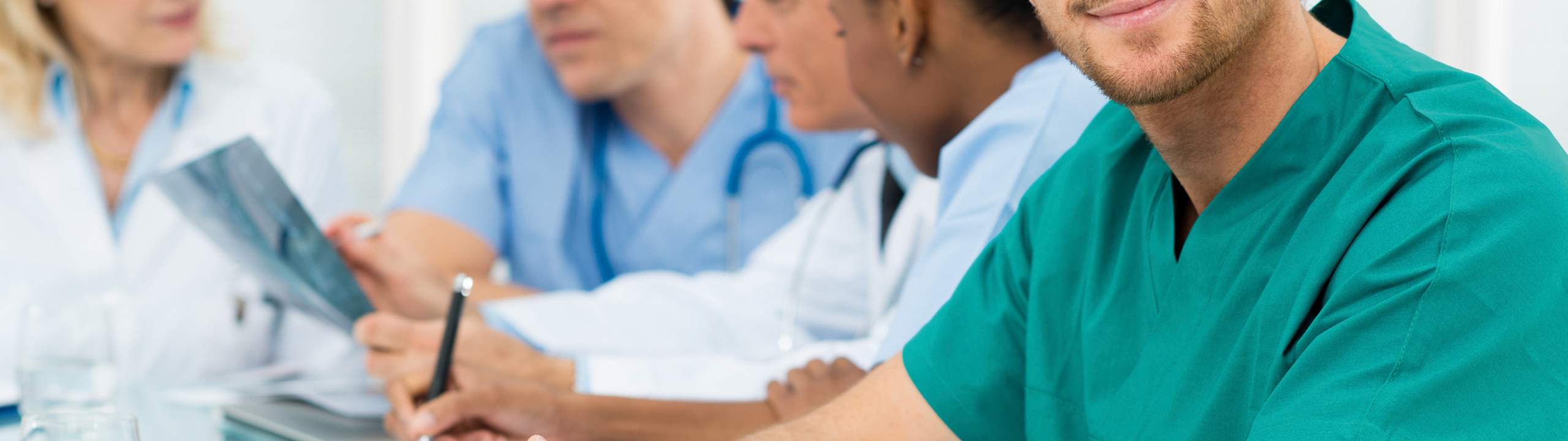 Ein Arzt in grüner Arbeitskleidung lächelt, während Kollegen in einem Besprechungsraum beraten und Unterlagen prüfen.