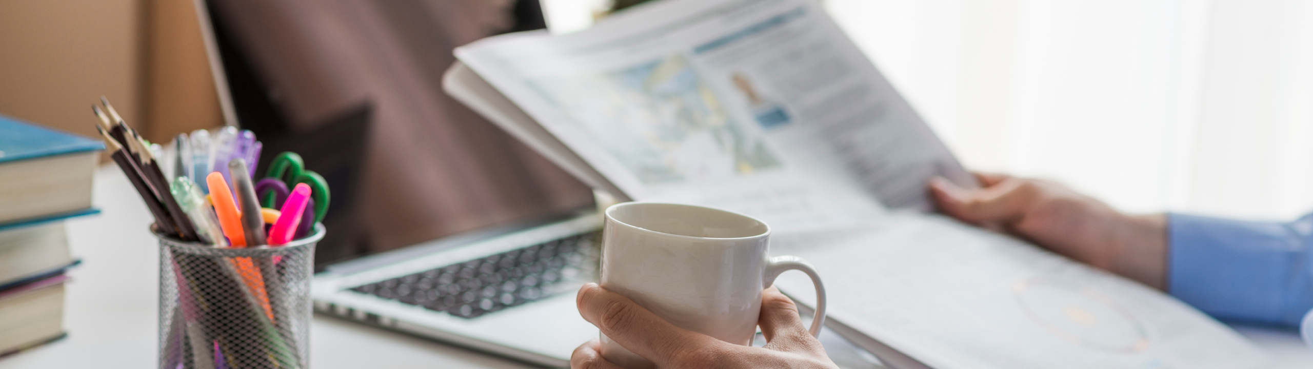 Eine Person hält eine Tasse Kaffee in der Hand und liest eine Zeitung am Schreibtisch mit Bürobedarf.